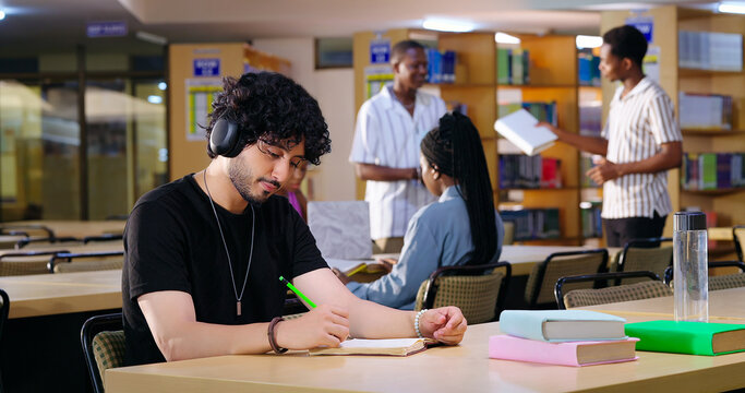 Indian Asian young adult gen z male man calm peace mind student sitting desk table listening music hold pencil write exam notes smart boy do self study read book prepare topics indoor library busy day