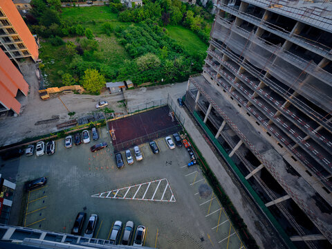 Aerial view of an urban courtyard with parking and cars beside a construction building with scaffolding, facade renovation and empty lot, greenery and driveway from high perspective.