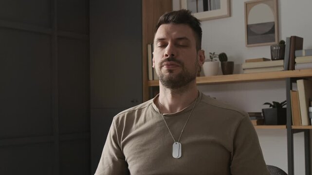 Chest-up view of Caucasian young bearded veteran soldier wearing identification tag deeply breathing with closed eyes during stress management therapy