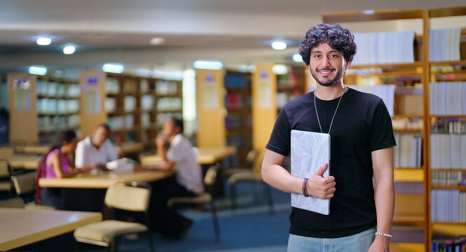 Indian young adult gen z man student standing proud hold new laptop look camera indoor library happy smiling boy guy staring cam do casual pose enjoy new joyful day time inside college