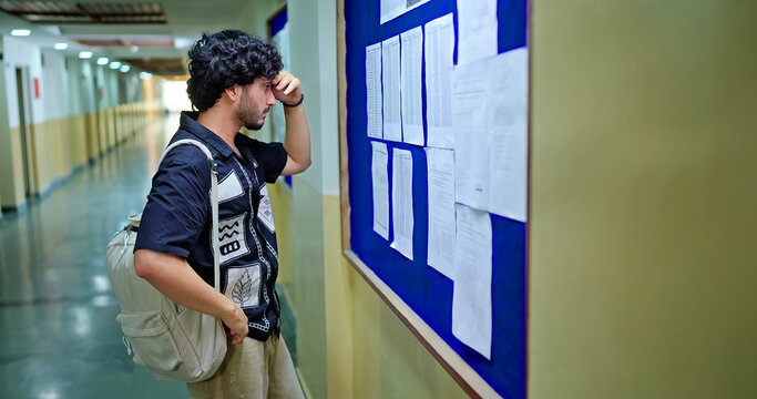 Indian young adult gen z male guy look notice board do check results fail in exam report feel bad campus area. Stress upset tensed teen pupil boy wear bag read new paper study name list time place