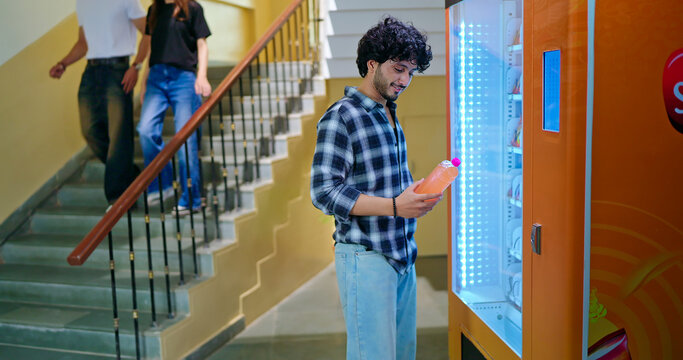 Indian happy young adult gen z smart male man use easy fast self service choose juice drink campus area. Lady female walk stairway thirsty boy take bottle enjoy break day time hallway