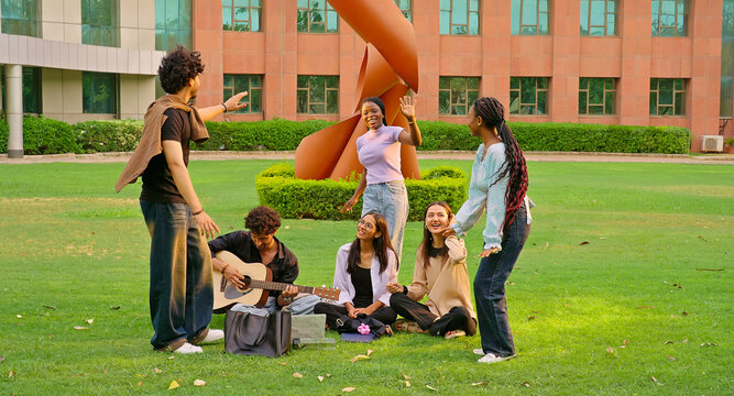 Indian young adult gen z male man hold guitar play new music lady woman sing song have fun joy. Happy pupil girl boy guy group spend day break time do funny dance enjoy relax mood at garden park place
