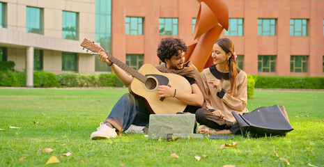 Indian young adult gen z male man hold guitar play music lyric lady woman sing song outdoor garden park place. Happy pupil girl boy pair sitting spend break time enjoy relax mood fun joy new day	