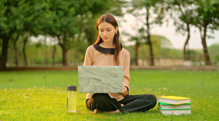 Fototapeta premium Indian young adult gen z busy lady female use laptop look screen check online new exam study notes garden park pupil girl sitting on grass do work read course email spend good break time day place