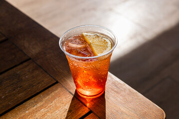 A refreshing alcohol drink in a clear plastic cup with ice cubes and a slice of orange. The drink is placed on a wooden table with soft lighting