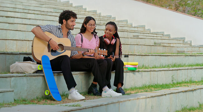 Indian young adult gen z male man sitting on stair hold guitar play lyric female sing song have fun joy place. Happy relax teen girl boy group spend good break time enjoy music mood new day stairway