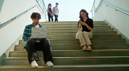 Indian young adult gen z busy male man use laptop look screen do study check online e work at campus place. Happy female sitting on stair hold mobile phone read notes enjoy new day spend break time