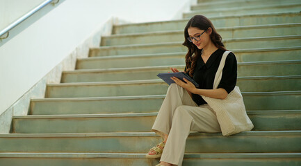 Indian young adult gen z lady woman use tab look screen do study exam check e learning read notes.. Teen girl wear glass sitting on stairs hold tablet causal enjoy new day spend break time place