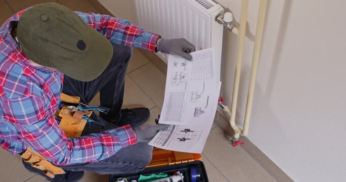 Professional plumber kneels by heating radiator reviewing schematic sheet. Male worker selects correct coupling from kit checking part code on chart during process