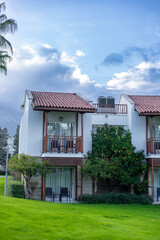 Two-story white building with a red-tiled roof, surrounded by green grass and palm trees. Cloudy sky in the background.