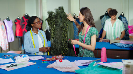 Indian young adult gen z two female wear tape around neck check dummy cloth make dress. Teen tailor girl pair talk speak share idea do paper work learn new course craft skill day at class room place