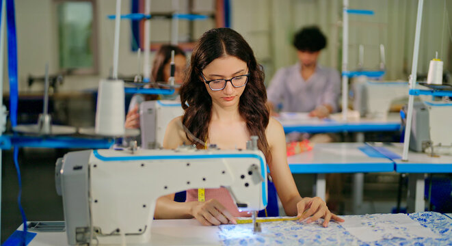 Indian young adult pupil gen z woman wear glasses tape around neck sitting class room learn new casual course good sewing machine. focused teen female girl do self craft skill indoor day college place - Powered by Adobe