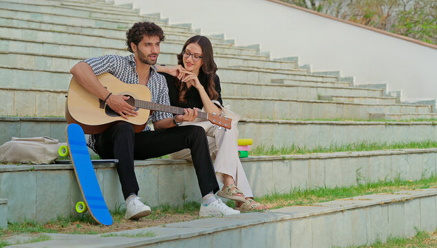 Indian young adult gen z female male man hold guitar play new lyric music sing song have fun joy stairway place. Happy relax teen girl boy guy sitting on stair enjoy good mood spend break day time