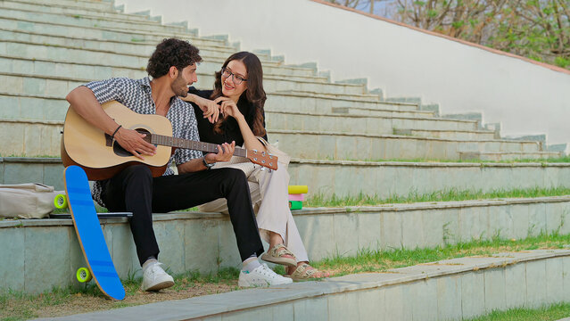 Indian young adult gen z female male man hold guitar play new lyric music sing song have fun joy stairway place. Happy relax teen girl boy guy sitting on stair enjoy good mood spend break day time
