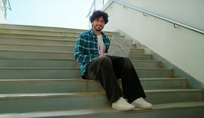 Indian young adult gen z male man use laptop look screen do study read email work check online e learning course break time. Happy pupil boy staring cam do casual pose enjoy new day outdoor place