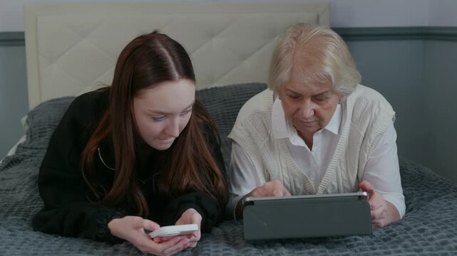 Elderly grandmother learning digital skills from supportive granddaughter, lying together on bed while using tablet and smartphone, bridging generational technology gap with love and patience