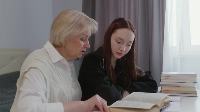 Senior woman guiding teenage granddaughter through textbook, sharing knowledge and creating supportive learning connection within home environment