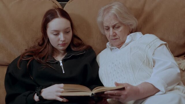 Elderly grandmother and young granddaughter lying closely on soft sofa, sharing intimate reading moment with deep familial connection and educational interaction
