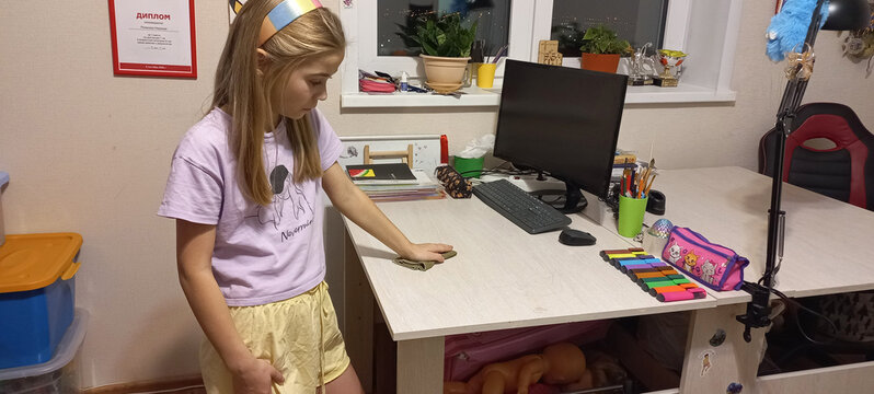A young girl wipes a table with office supplies with a rag in a home office - Powered by Adobe