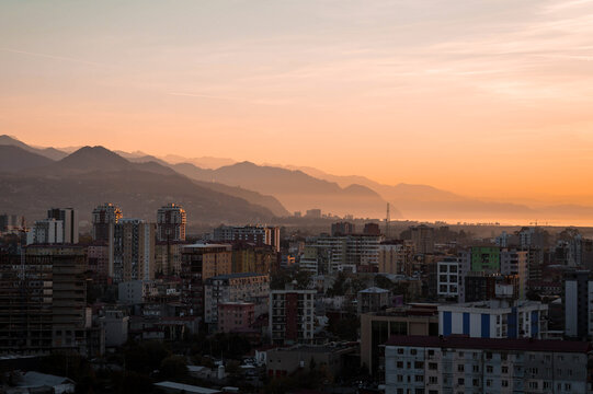 Sunset view over Batumi with silhouettes of Gonio and layered mountain ridges. Warm golden light illuminates the city skyline, sea haze and distant hills, creating a peaceful evening atmosphere.