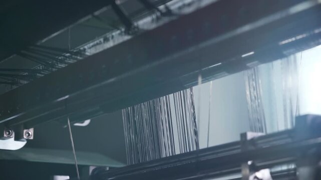 Threads pass through the structure of an industrial weaving machine while soft horizontal anamorphic flares highlight the metal frame and the motion of textile production