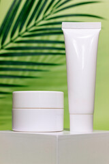 close up of empty white jar and tube of cosmetic cream on a white square shaped podium against light green background with soft shadow of leaves