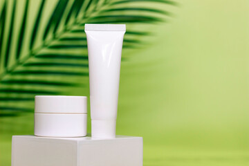 empty white jar and tube of cosmetic cream on a white square shaped podium against light green background with soft shadow of leaves and copy space for text