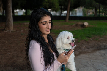 Woman holding maltese dog using smartphone in park