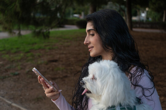 Woman holding maltese dog using smartphone in park
