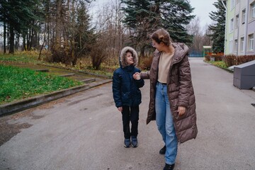 Obraz premium A young mother picks up her son from school, walking home together with joyful smiles. Authentic moment of family love, education, and daily parenting routine
