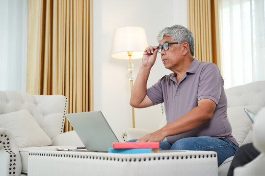 Senior man working on a laptop in the living room, Elderly man adjusting glasses while using a computer, Older man focusing on online tasks at home