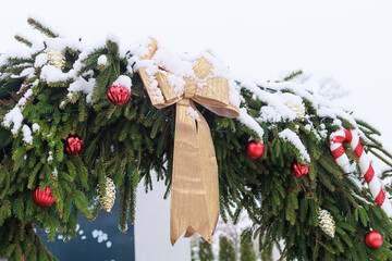 Outdoor christmas garland with snow, evergreen branches, red baubles and a large bow made of golden ribbon. Concept of celebration, city decoration, postcard, banner and winter design.