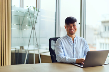 Fototapeta premium Asian Man in a Bright Workspace Smiling While Working on Laptop in Modern Office