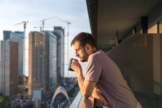 A man standing on a balcony during golden hour, drinking coffee and looking at the city skyline. Modern urban lifestyle, peaceful morning atmosphere, high-rise buildings and warm sunlight. - Powered by Adobe