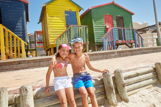 Portrait, brother and sister at beach with hug, bonding together and summer holiday by colorful huts. Happy, children and siblings with embrace at ocean for connection, vacation and weekend break. - Powered by Adobe