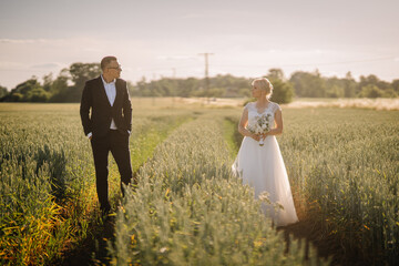 Fototapeta premium Bride in elegant white gown holding bouquet stands in a wheat field, while groom in black suit gazes at her, capturing a romantic outdoor wedding moment with nature's beauty