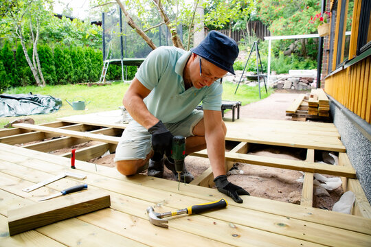 Homeowner using a power drill to install wooden deck planks during a backyard renovation, capturing realistic DIY, woodworking, outdoor construction.