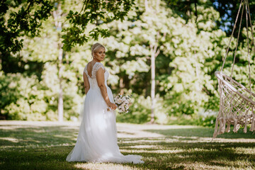 Fototapeta premium Bride in elegant white gown stands in lush green park, holding bouquet, with sunlight filtering through trees, creating a romantic and serene atmosphere for wedding celebration