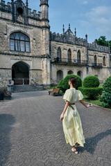 Naklejka premium Young woman in a pale yellow dress walks across a gravel courtyard toward a grand historic castle, capturing a serene travel moment with architectural charm and soft light.
