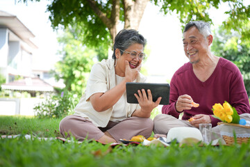 Senior Couple Enjoying A Picnic While Watching A Tablet Together, Happy Elderly Couple Relaxing Outdoors And Sharing A Laugh, Older Couple Having A Picnic And Watching Content On A Tablet