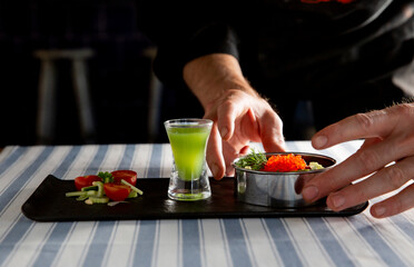 Chef hands serving an elegant gourmet appetizer with red caviar and vegetables. Paired with a vibrant green shooter cocktail.