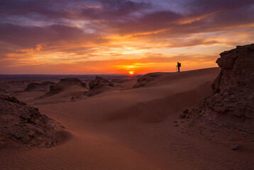 Silhouette of hiker on desert dune ridge at sunset. Vivid orange and purple sky over vast sand landscape and rocky formations. Solitude and adventure in natural wilderness.