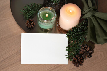Blank paper card mockup, candles burning, pine tree branches and cones on neutral beige wooden table background. Aesthetic winter postcard or invitation design template with copy space, top view