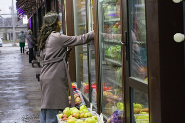 young woman in a winter coat at an outdoor market, standing by the cashier, handing over money for fresh fruits in a crate. Perfect for articles about groceries, payments, or everyday city life.