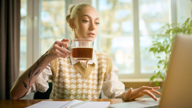 Employee holding coffee while working at laptop in bright home office. Concept of workday routines, caffeine boost, remote productivity, coworking lifestyle, focused morning energy.
