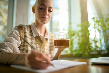 Woman holding coffee while writing notes in bright office. Concept of structured workflow, productivity habits, office lifestyle, and beverage-friendly creative planning.