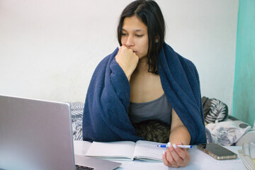 Indian student in a blue blanket sits at a desk with a laptop, open notebook, and smartphone, watching a lecture online. Her cat is nestled in her lap. Perfect for study, at-home education topics.
