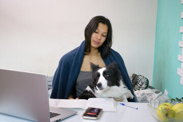 Young woman in a blue blanket studies at a desk using a laptop, with a dog on her lap, notebook, phone and fruit on the table. Good for distance learning, pets, or daily routines at home.