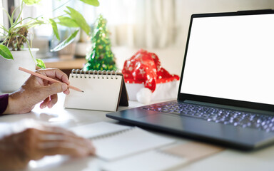 close up shot, hand holding pencil taking note on blank calendar working with laptop, red hat, christmas tree for decoration on desk, christmas holidays concept